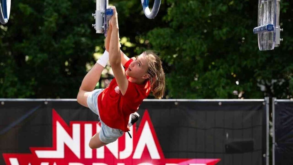 Person klettert bei Ninja Warrior-Wettbewerb, zeigt Sportlichkeit und Geschicklichkeit vor grünem Hintergrund.