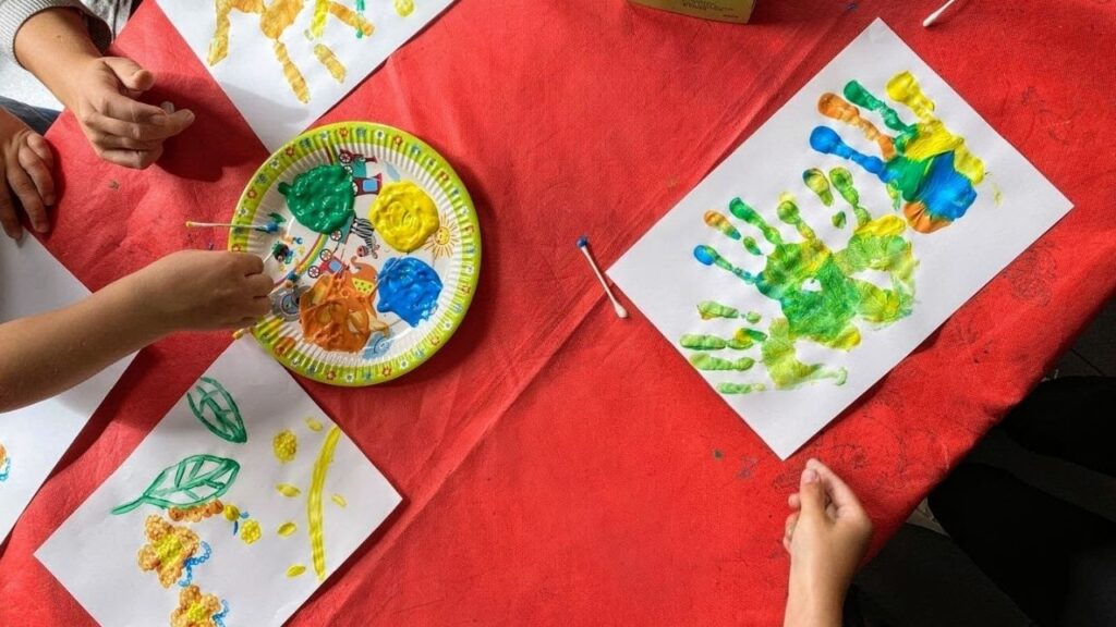 Kinder malen bunte Handabdrücke und Bilder mit Fingerfarben auf Papier. Kreatives Basteln auf rotem Tisch.