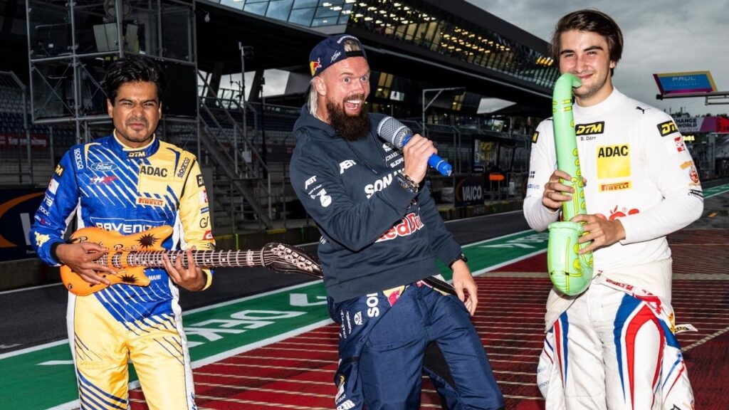 Rennfahrer mit Spielzeug-Gitarre und Mikrofon posieren humorvoll auf der Rennstrecke.