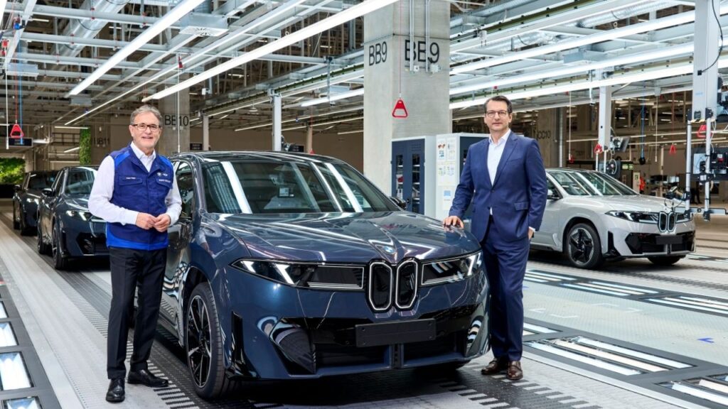 BMW-Fabrik, zwei Männer posieren neben einem neuen Auto auf der Produktionslinie, moderne Fertigung im Hintergrund.
