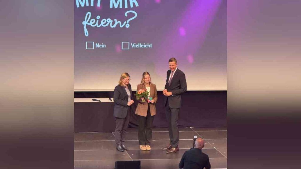 Drei Personen auf der Bühne, Frau in der Mitte mit Blumenstrauß, Text Mit mir feiern? im Hintergrund.