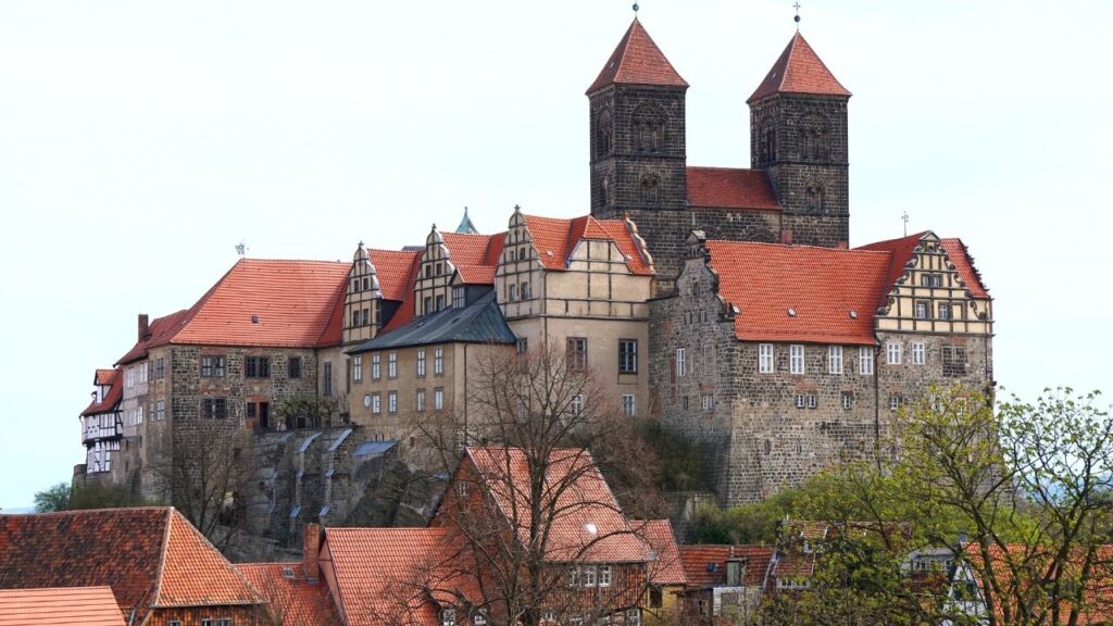 Mittelalterliche Burg mit roten Dächern, imposante Steinwände und Türme, umgeben von Bäumen bei klarem Himmel.