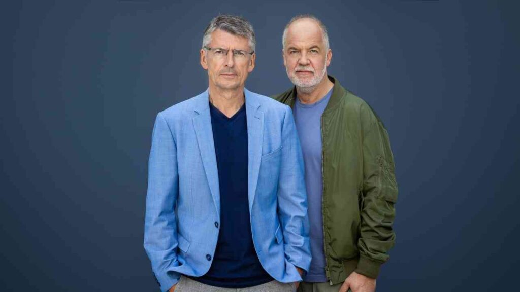 Zwei Männer vor blauem Hintergrund, einer im blauen Anzug, der andere in grüner Jacke, beide ernst blickend.