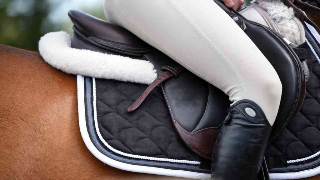 Reiter in heller Reithose auf Sattel, Detailansicht mit schwarzer Schabracke und Stiefel.