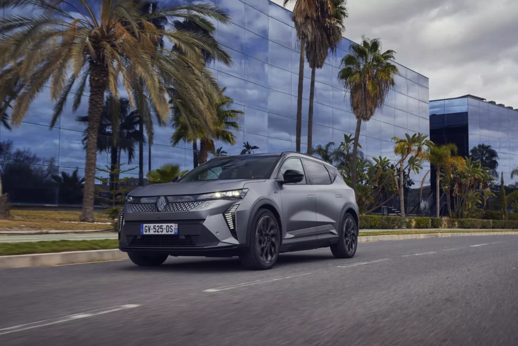 Grauer SUV fährt auf Straße, umgeben von Palmen, vor einem modernen Glasgebäude bei bewölktem Himmel.
