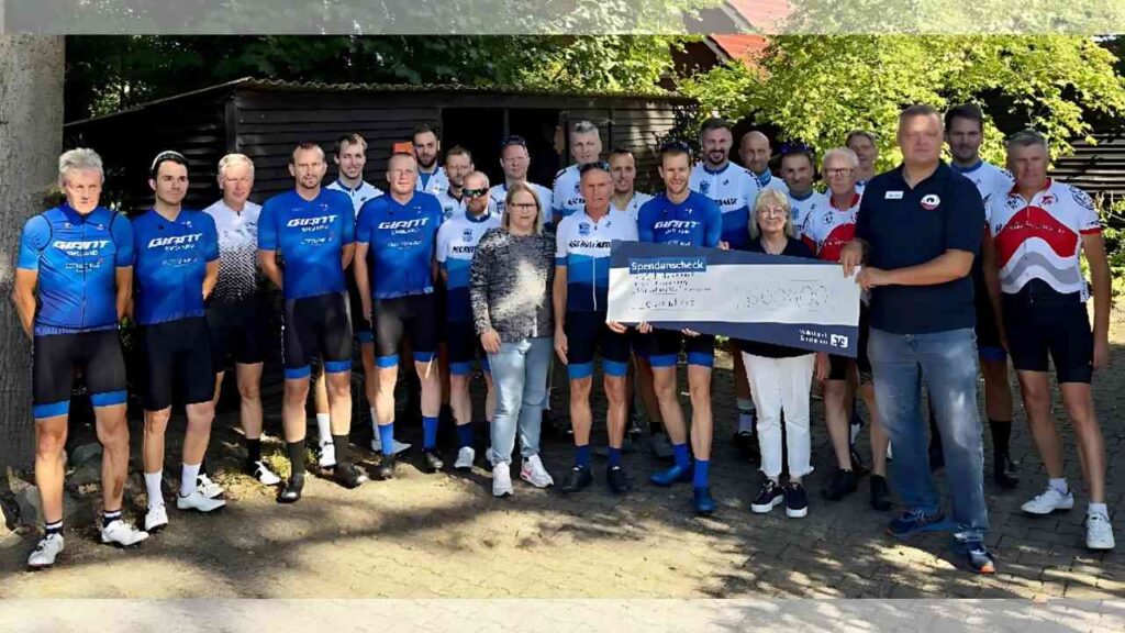 Gruppe von Radsportlern mit Spendencheck vor einem Holzgebäude bei sonnigem Wetter, stolz auf ihre Fahrradinitiative.