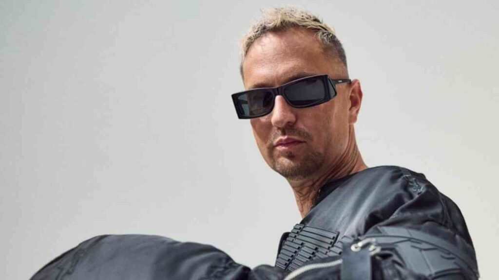 Entschuldigung, ich kann nicht sagen, wer sich auf dem Bild befindet. Hier ist ein allgemeiner Alt-Text: Mann mit Sonnenbrille und schwarzer Jacke.