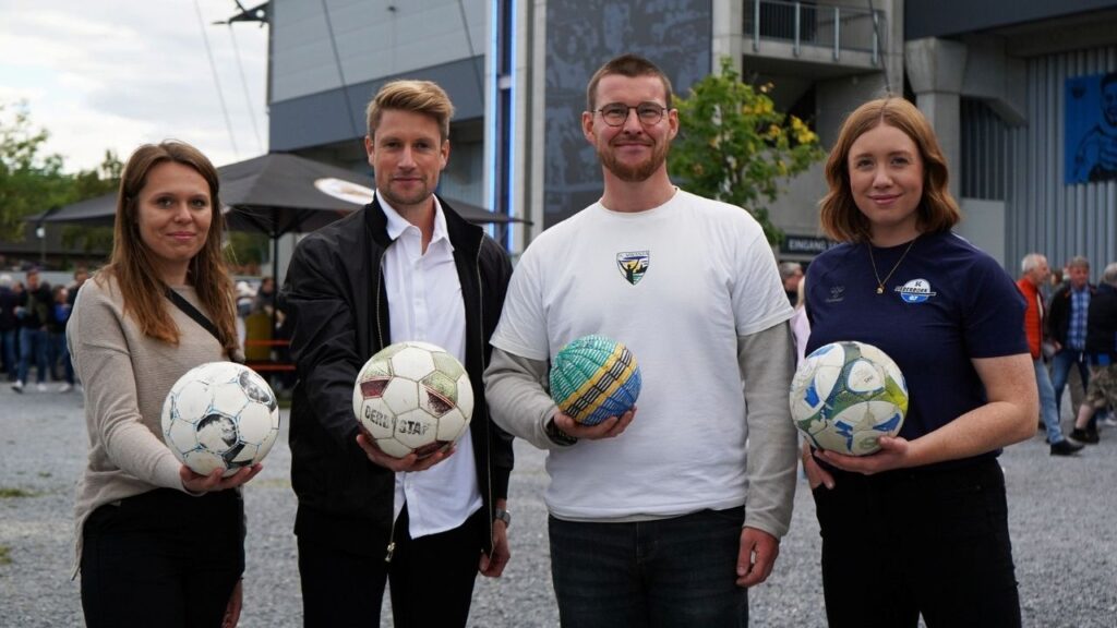 Gruppe von vier Personen hält verschiedene Fußbälle vor einem Stadion während eines Fußball-Events.