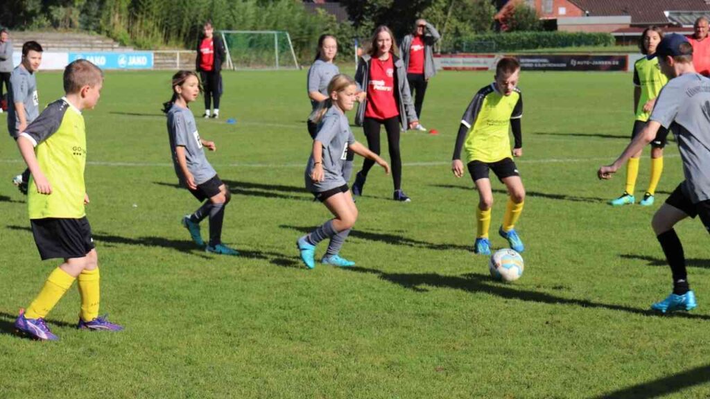 Kinder spielen Fußball auf grünem Spielfeld bei sonnigem Wetter, überwacht von Trainern im Hintergrund.