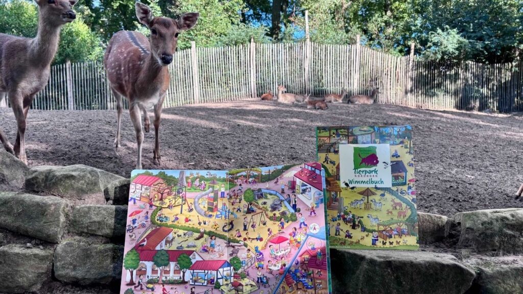 Rehe im Zoo vor einem Wimmelbuch mit Tierpark-Motiven, umgeben von Bäumen und einem Zaun im Hintergrund.
