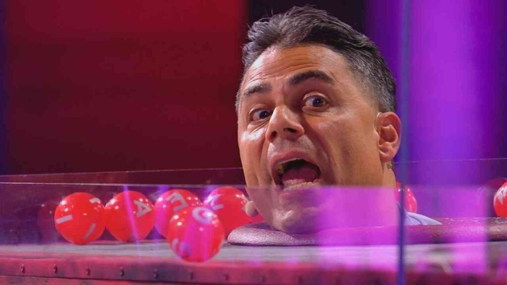 Person schaut aus Plexiglas-Box, umgeben von roten Bällen, in bunter TV-Show. Spannung und Überraschung sichtbar.