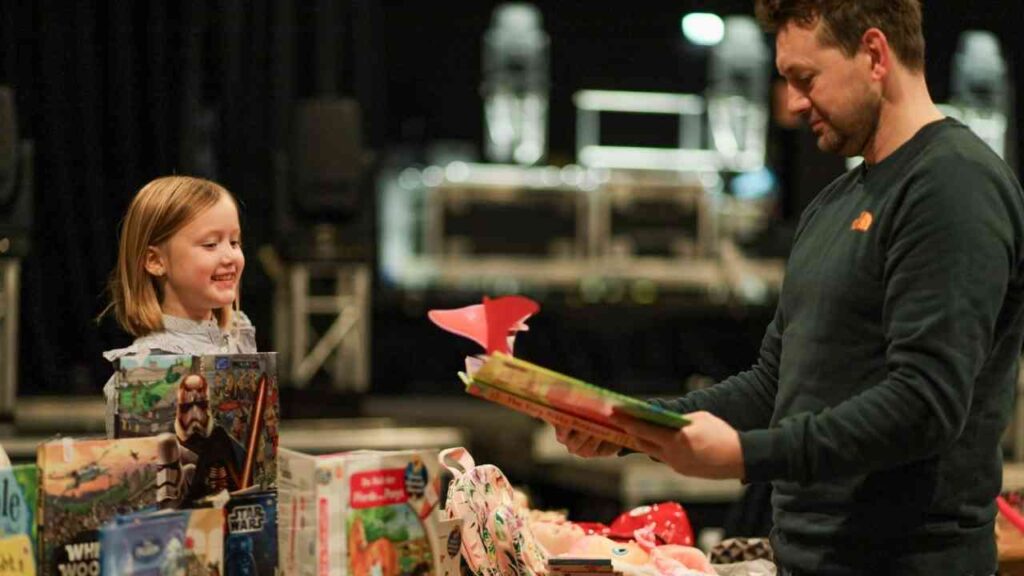 Erwachsener zeigt Kind Bücher auf einem Tisch, umgeben von Spielzeug in einem dunklen Raum.