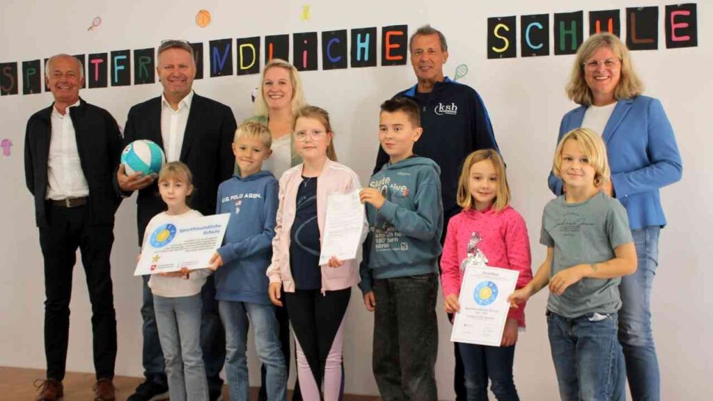 Gruppe von Erwachsenen und Kindern bei Verleihung eines Zertifikats für eine sportfreundliche Schule, ein Ball wird gezeigt.