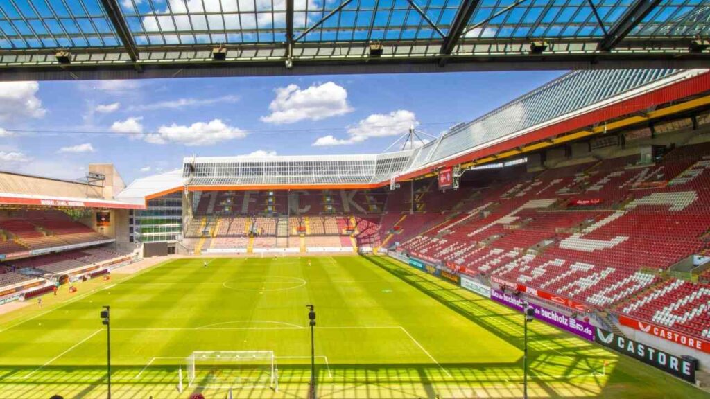 Leeres Fußballstadion bei Sonnenschein mit grünem Spielfeld und überdachten Tribünen, ideal für Sportveranstaltungen.