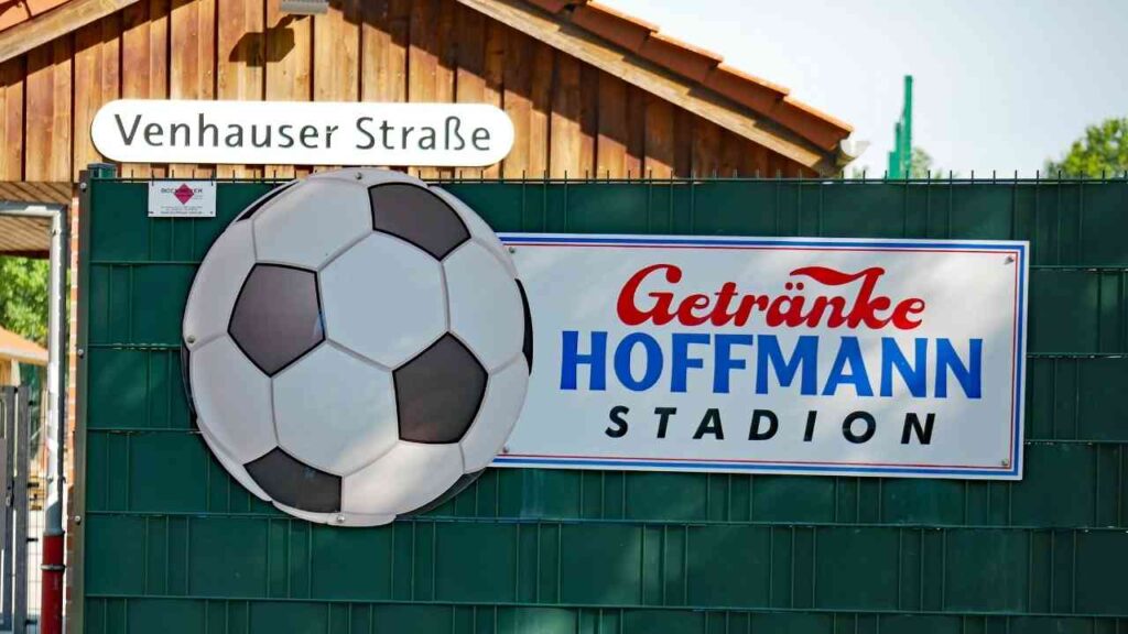 Schild des Getränke Hoffmann Stadions mit großem Fußball, Venhauser Straße im Hintergrund, Sonnenschein.