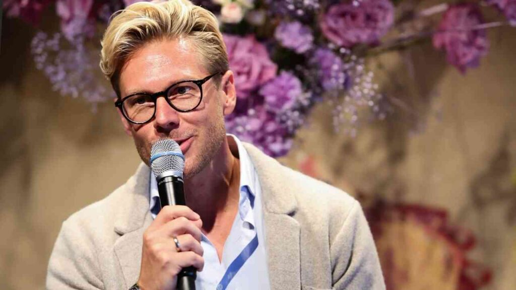 Person mit Mikrofon und Brille spricht vor lila Blumenhintergrund.