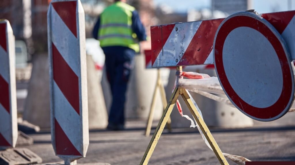 Straßenbaustelle mit Absperrungen, Verkehrskegel und Arbeiter in Warnweste im Hintergrund bei sonnigem Wetter.