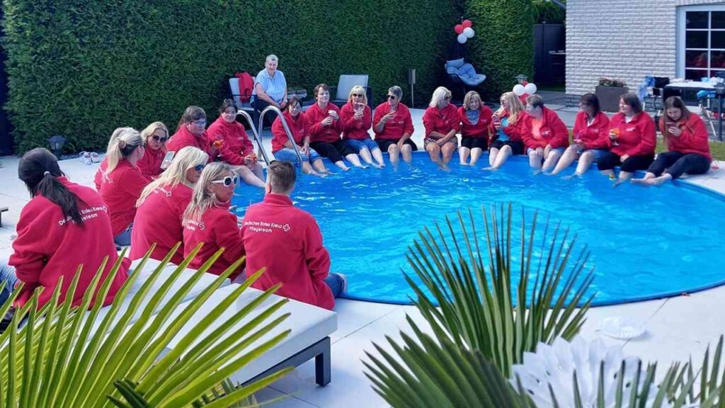 Gruppe in roten Jacken sitzt am Pool, entspannt und lächelt, sonniger Tag, Hecke im Hintergrund, Sommerfest-Atmosphäre.
