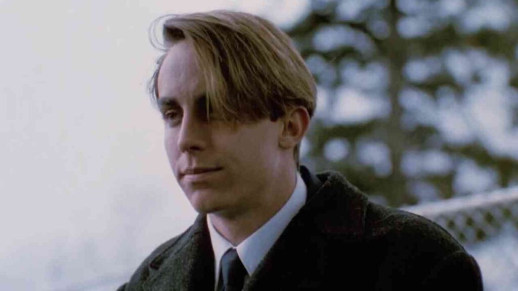 Junger Mann mit mittellangen Haaren im Anzug schaut nachdenklich seitwärts, vor unscharfer Naturkulisse.