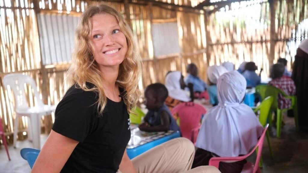 Person lächelt in einer Schulklasse, Kinder sitzen auf bunten Stühlen in einem Raum mit Bambuswänden.