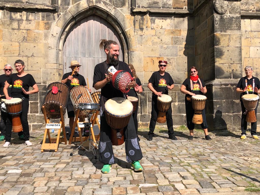 Gruppe von Musikern spielt traditionelle Trommeln im Freien vor einer alten Steinmauer, sonniger Tag.
