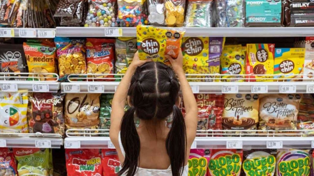 Mädchen greift nach Süßigkeiten im Supermarktregal, umgeben von bunten Snack-Verpackungen.