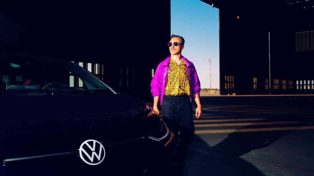 Person in lila Jacke und Sonnenbrille neben VW-Auto im Sonnenschein, Flughafenhangar im Hintergrund.