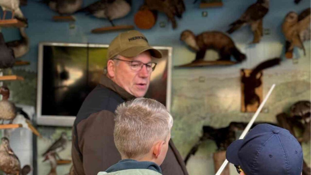 Mann erklärt Kindern Tiere in einem Museum, umgeben von ausgestopften Exponaten und einem Fernseher im Hintergrund.