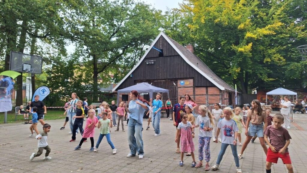Kinder tanzen fröhlich bei einem Freiluft-Event vor einer rustikalen Scheune, umgeben von grünen Bäumen.