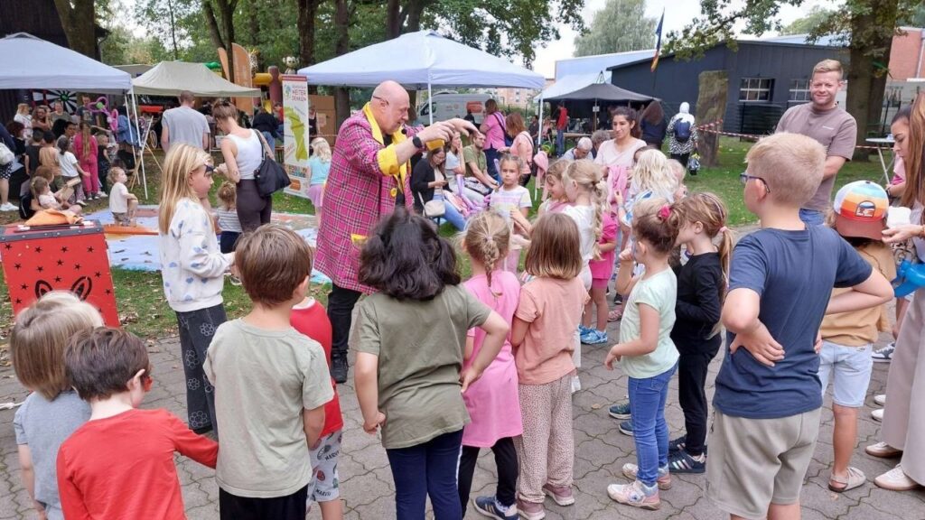 Kinder im Kreis um einen Künstler bei einem bunten Outdoor-Sommerfest mit Ständen und Zuschauern.