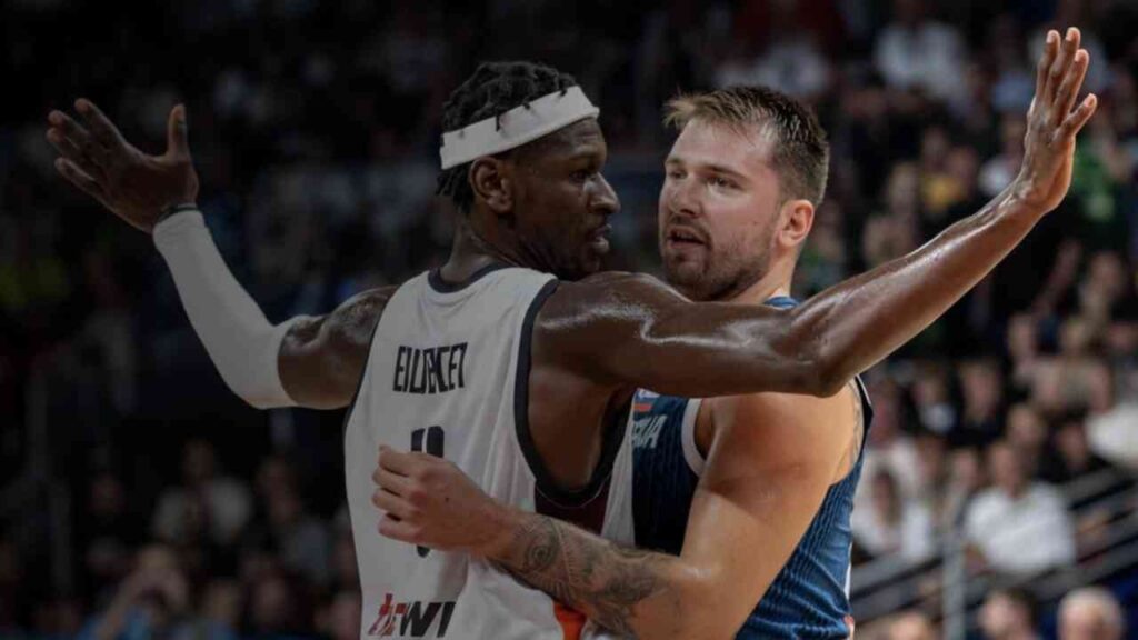 Basketballspieler in intensivem Zweikampf auf dem Spielfeld, einer in weiß, der andere in blau, vor einer unscharfen Zuschauermenge.