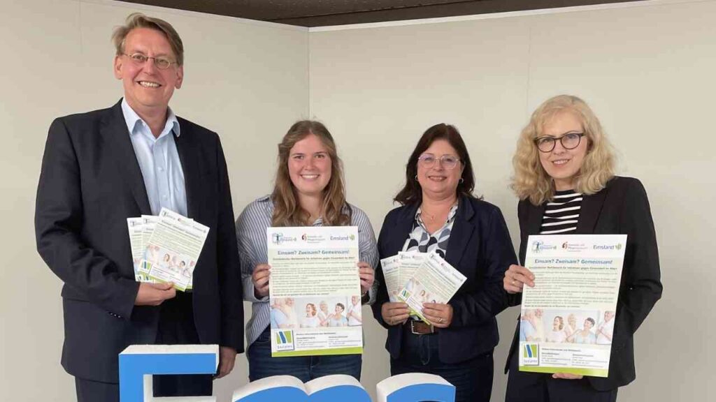 Gruppe von vier Personen präsentiert Informationsbroschüren für ein soziales Projekt, stehend vor einem Emsland-Logo.