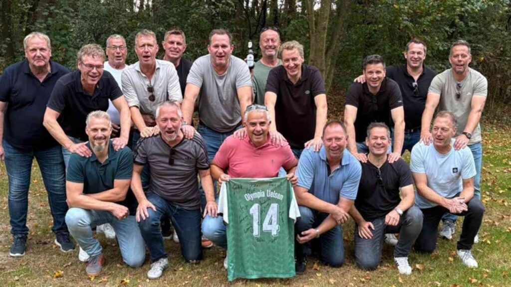 Männergruppe im Freien mit Trikot von Olympia Uelsen, Nummer 14, grünem Hintergrund, Herbststimmung, lächelnd.