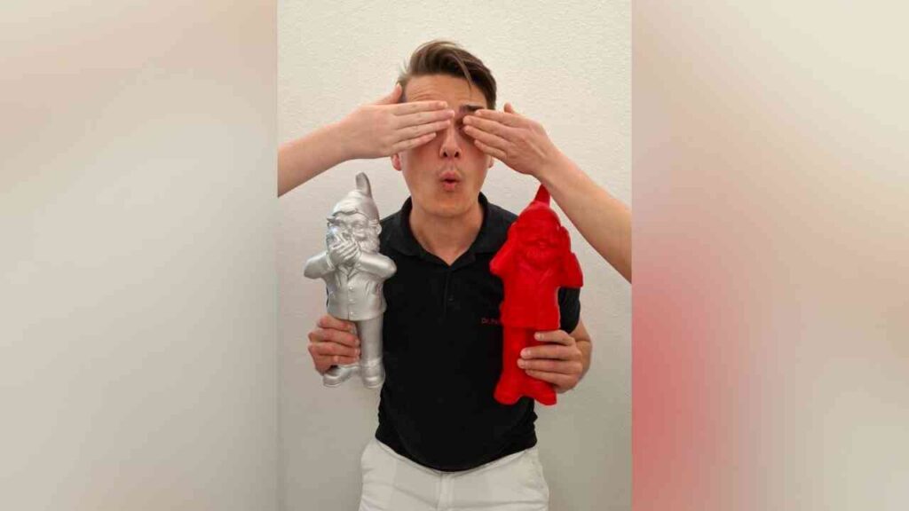 Person hält rote und silberne Gartenzwerge, während Hände die Augen verdecken. Überraschungsausdruck im Gesicht.