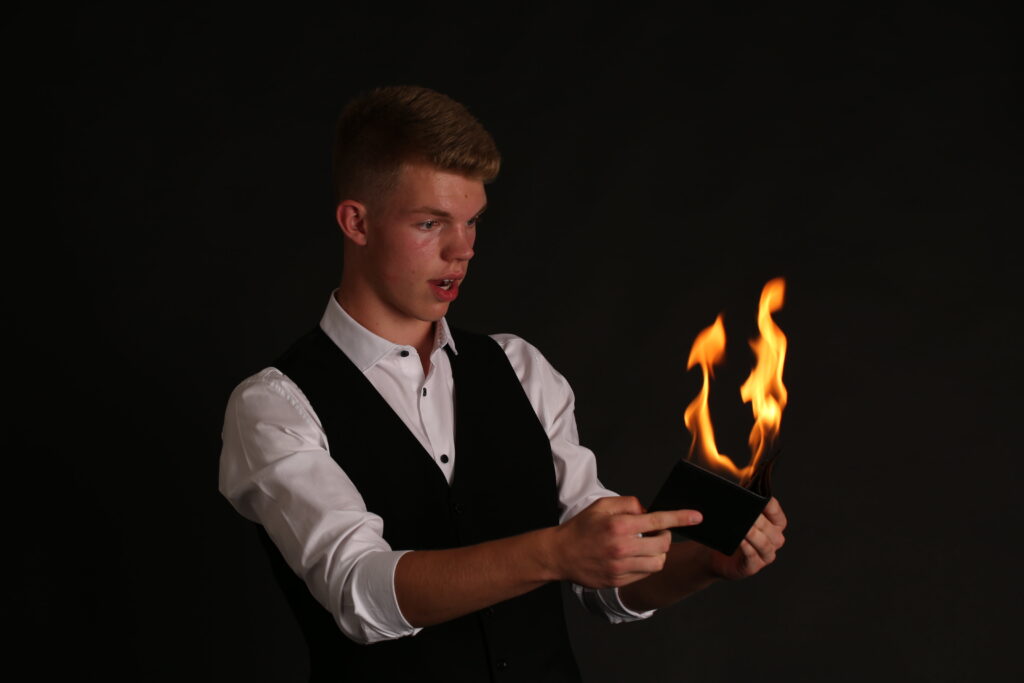 Mann im Anzug hält brennendes Portemonnaie, überrascht. Magie- oder Trickeffekt in dunklem Studiohintergrund.
