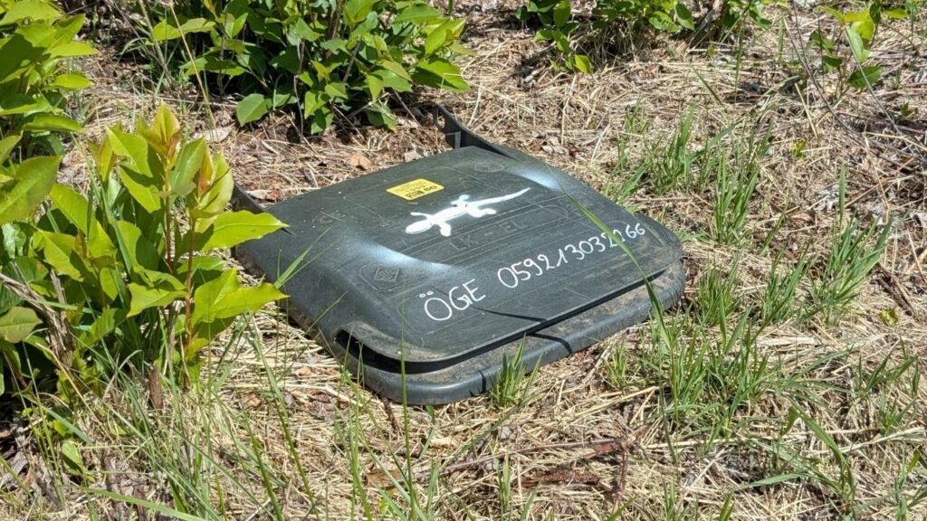 Schwarzes Kästchen im Gras mit Krokodil-Logo und Aufschrift ÖGE 059213031266, umgeben von Pflanzen.