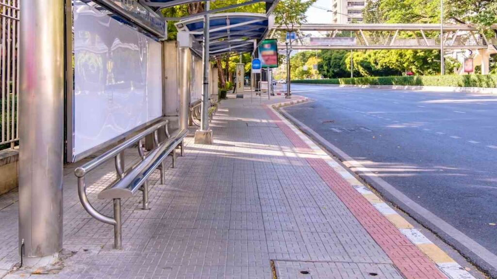 Leere Bushaltestelle an einer sonnigen Straße, umgeben von Bäumen und Gehweg, mit Überdachung und Sitzbank.