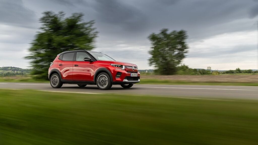 Roter SUV fährt auf einer Landstraße, während sich im Hintergrund Bäume und Felder erstrecken. Dynamische Fahraufnahme.