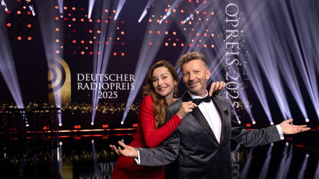 Moderatorenduo beim Deutschen Radiopreis 2025 auf Bühne mit Scheinwerfern im Hintergrund. Elegante Abendgarderobe.