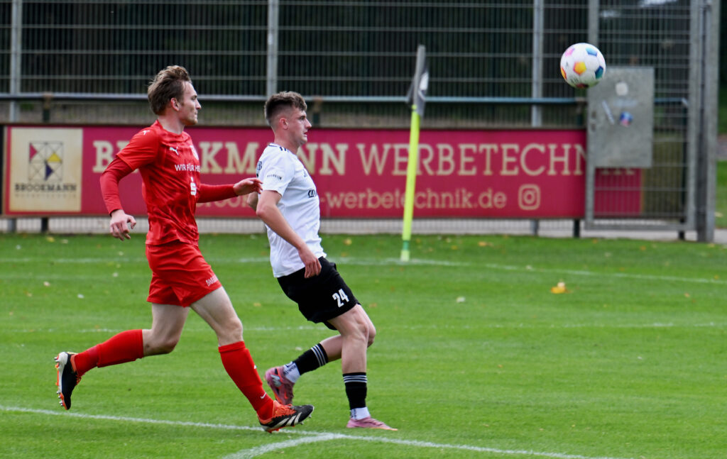 Zwei Fußballspieler im Spiel, einer in rotem Trikot, der andere in weißem, laufen zum Ball auf grünem Feld.