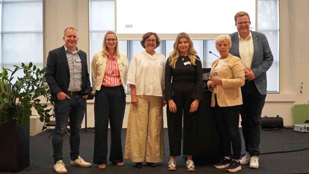 Gruppe von sechs Menschen posiert lächelnd bei einer Veranstaltung in einem modernen Konferenzraum.