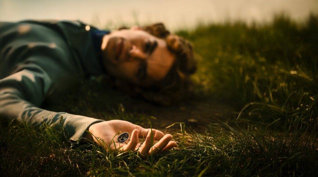 Junger Mann mit lockigem Haar, der auf dem Gras liegt und eine Uhr in der Hand hält.