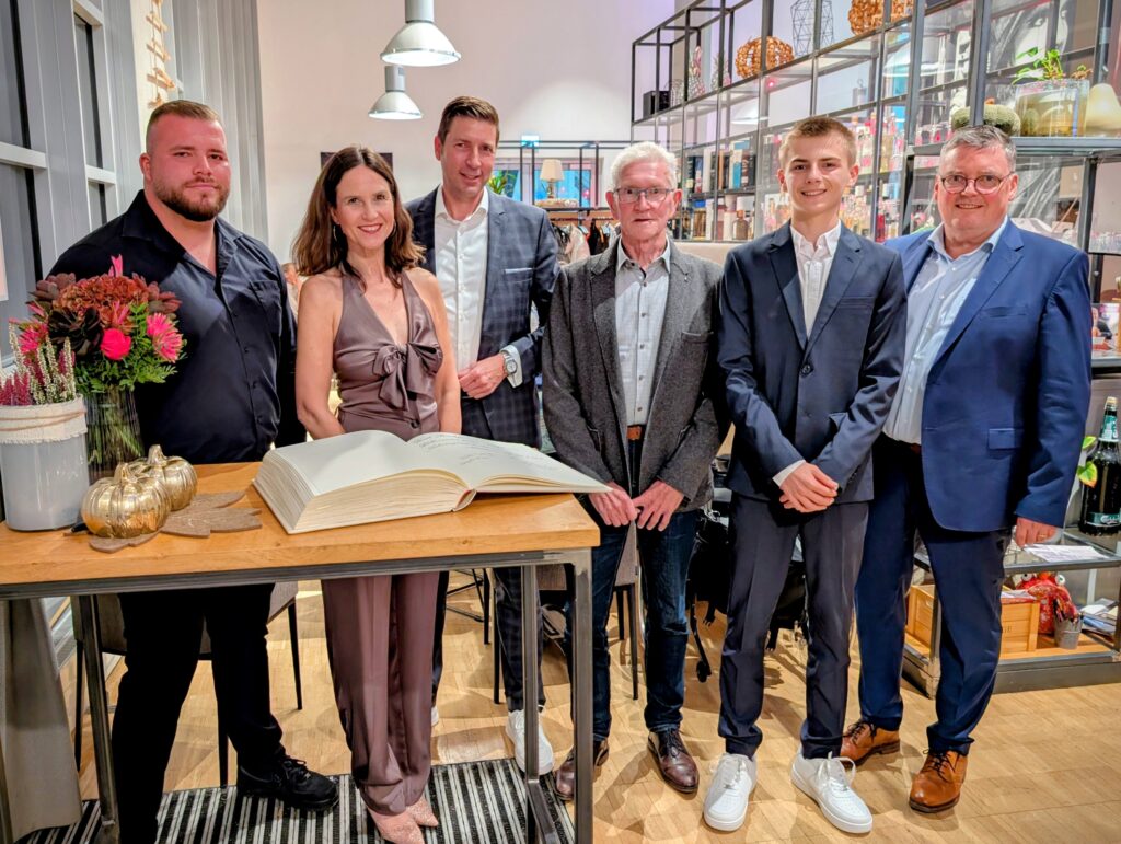 Sechs Personen posieren für ein Gruppenfoto in einem Innenraum mit einem großen Buch auf einem Tisch.