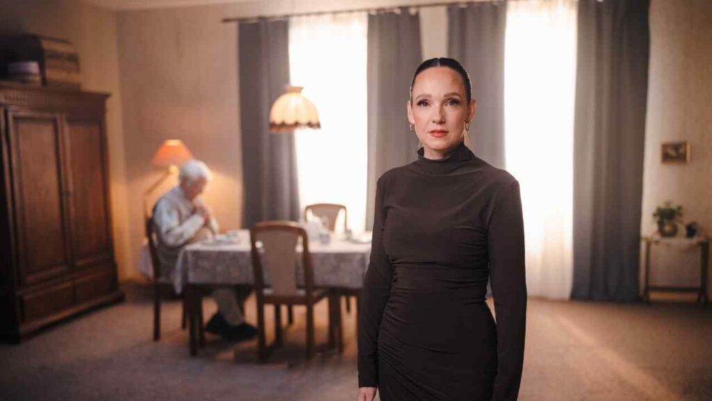 Frau in einem eleganten, dunklen Kleid steht in einem Raum mit einem Tisch im Hintergrund, an dem eine ältere Person sitzt.