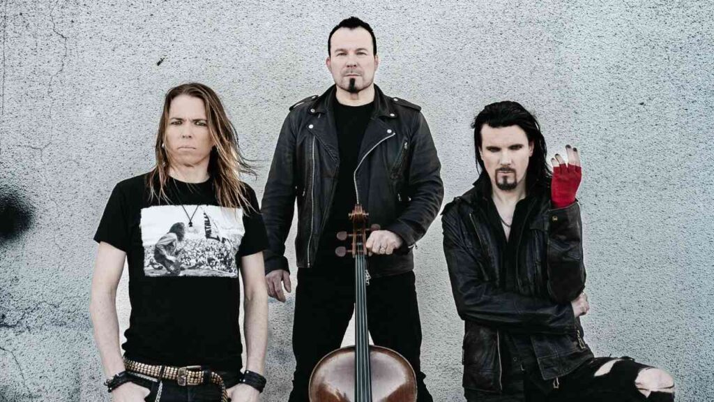 Drei Musiker posieren vor einer grauen Wand, einer hält ein Cello, während die anderen in lässiger Kleidung stehen.