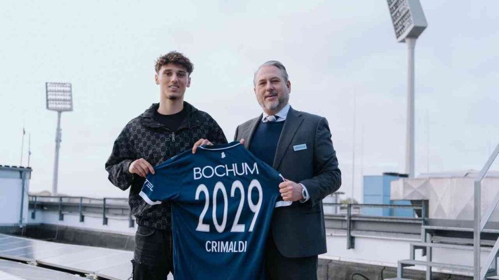 Alessandro Crimaldi hält ein Fußballtrikot mit der Aufschrift 'Bochum 2029' in der Hand, während er von einem Mann im Anzug begleitet wird.