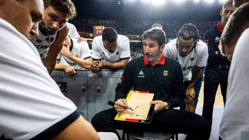 Basketballtrainer Alex Mumbru, der während einer Besprechung auf einem Notizblock schreibt, umgeben von Spielern.