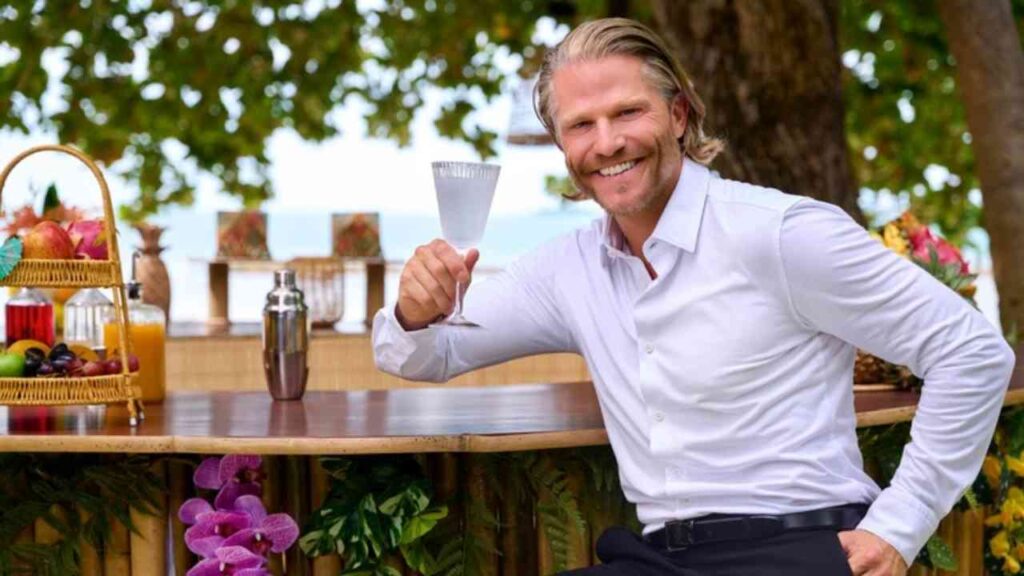 Mann mit einem Cocktail in der Hand, der an einer Bar im Freien sitzt und lächelt.