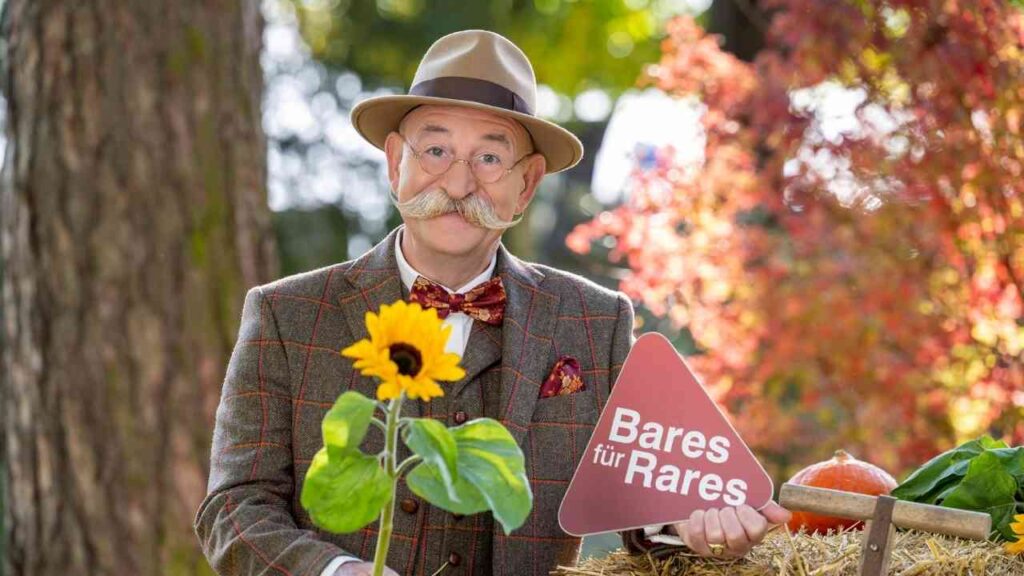 Mann mit Schnurrbart und Hut hält ein Schild mit der Aufschrift 'Bares für Rares' und eine Sonnenblume in der Hand.