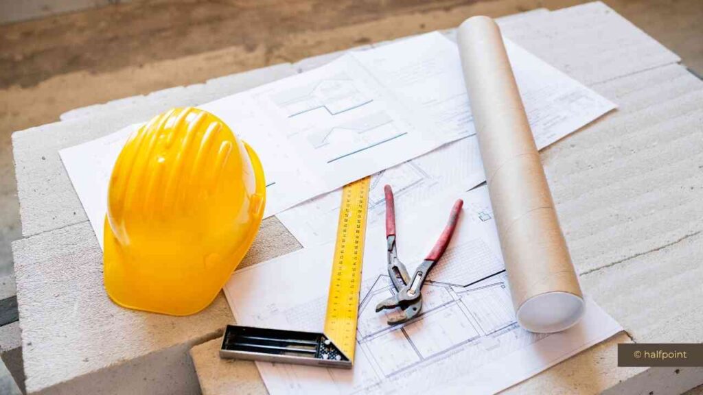 Gelbe Sicherheitshelmkappe, Baupläne, Zollstock und Zange auf einem Baustellenuntergrund.